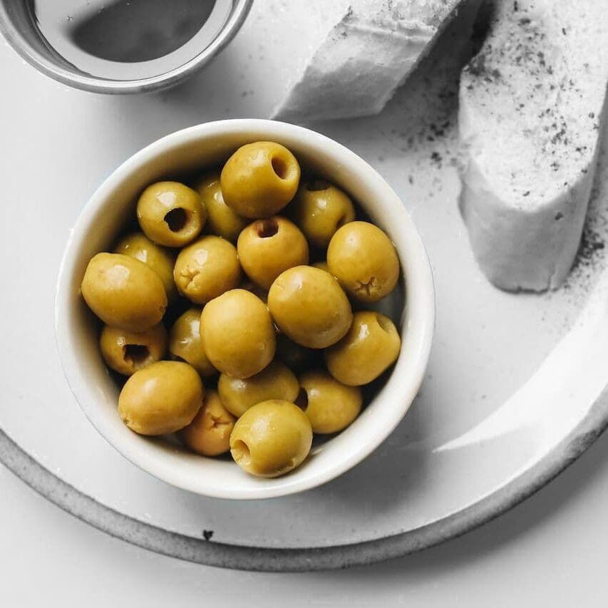 A bowl of olives on a tray with cheese.